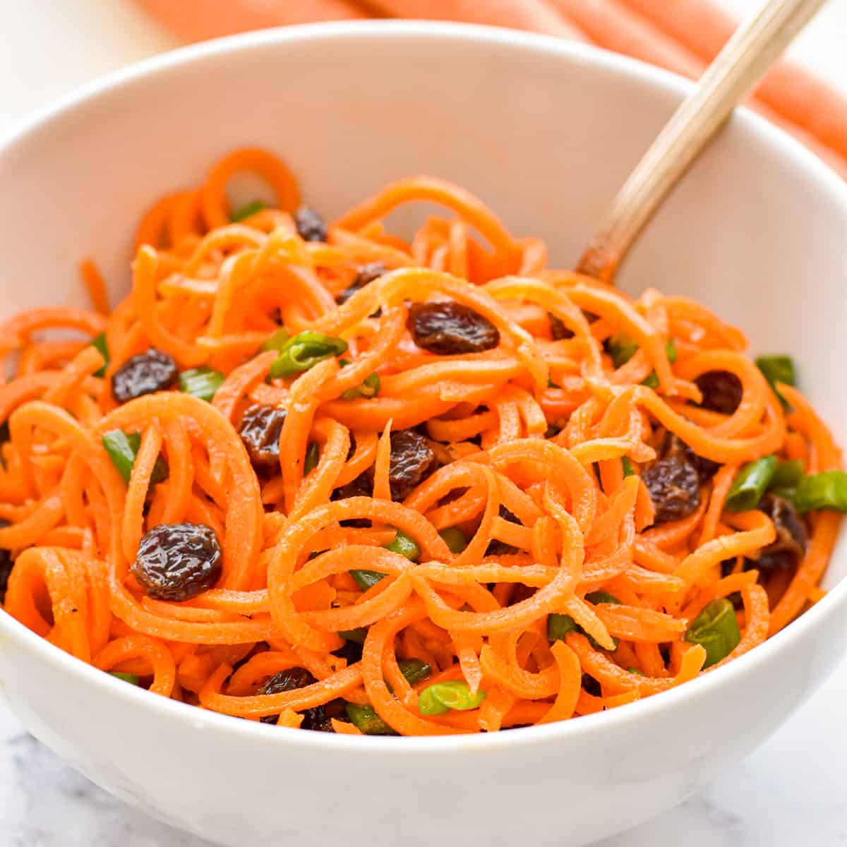 carrot slaw with spiralized carrots raisins and green onions in a white bowl with a spoon