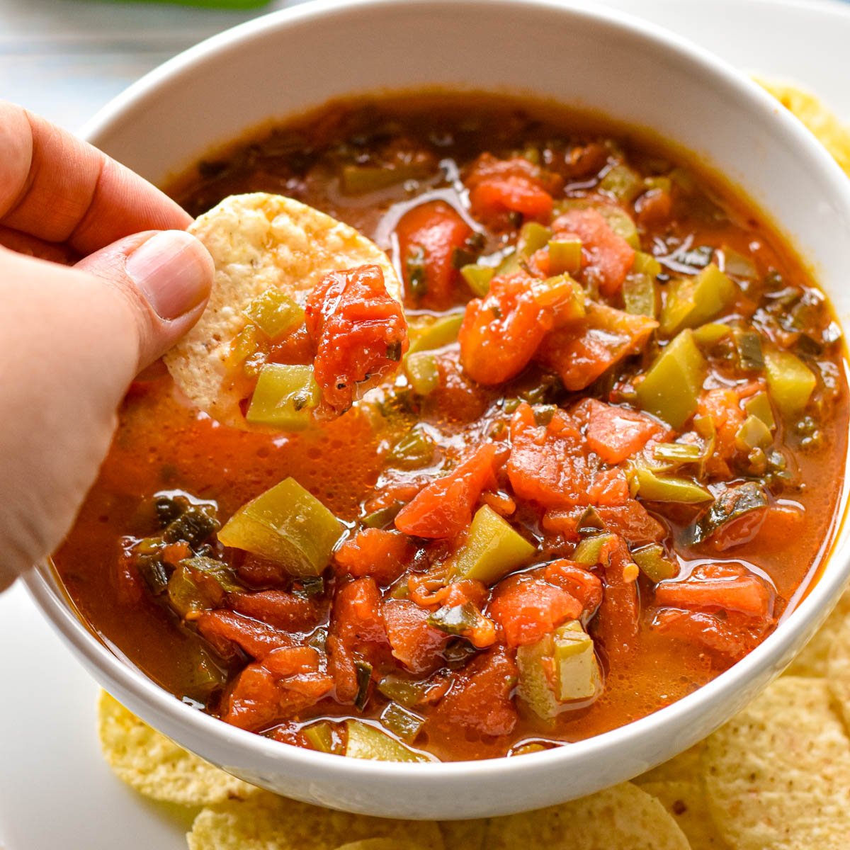 dipping a tortilla chip into low fodmap salsa in a white bowl surrounded by chips