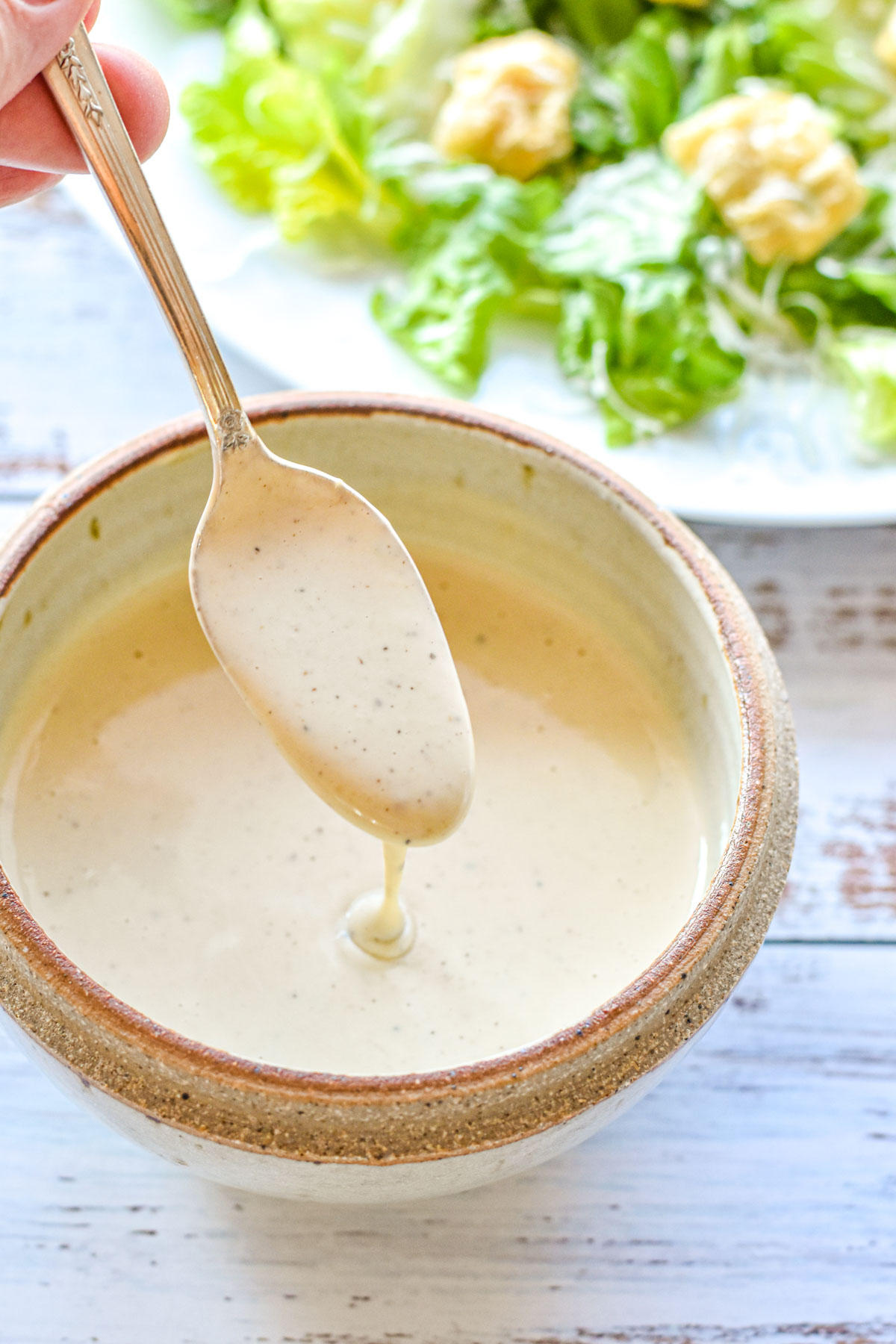 low fodmap Caesar dressing pouring out of a spoon into a pottery bowl with a salad in the background
