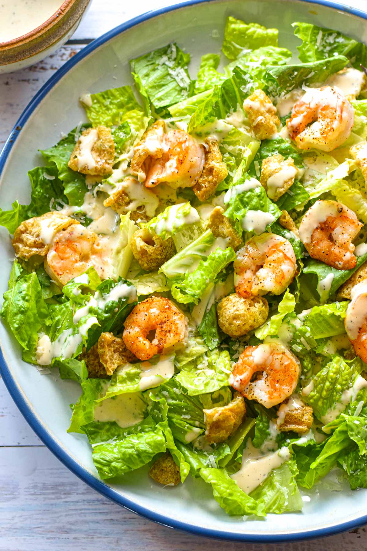 close up shot of low fodmap shrimp Caesar salad on a blue and white plate in front of a pottery bowl of low FODMAP Caesar dressing