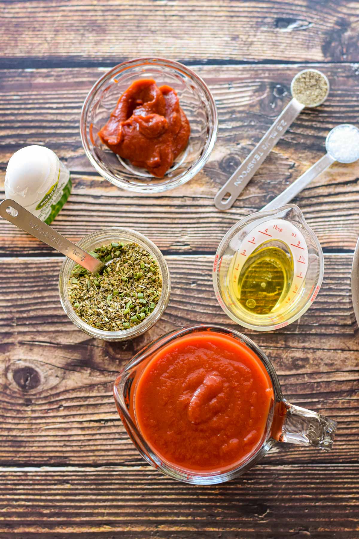 ingredinets of low FODMAP keto pizza sauce on a wooden background including strained tomatoes, garlic-infused olive oil, italian seasoning, salt, pepper, tomato paste, and stevia