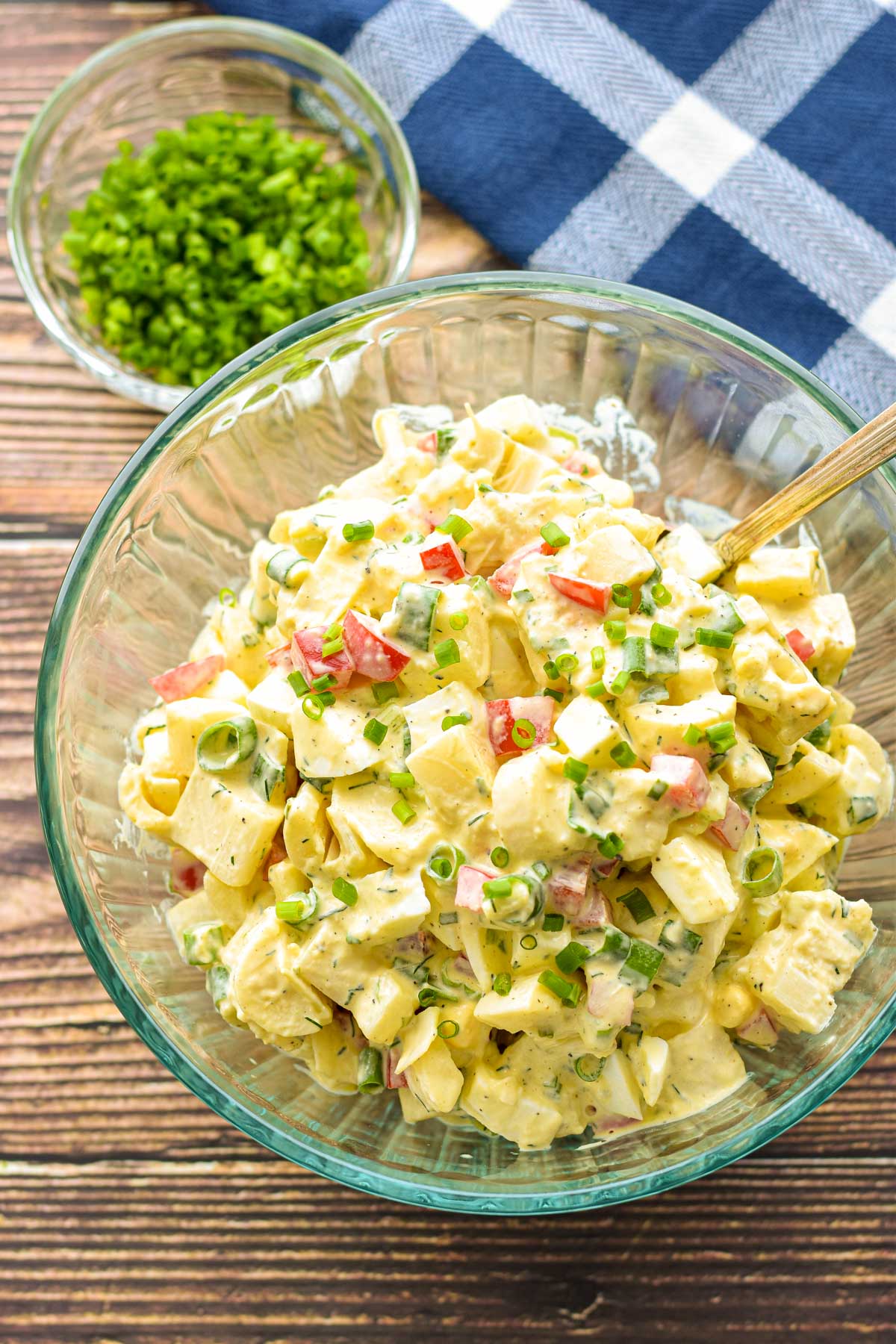 overhead shot of low fodmap keto faux potato salad in a clear bowl with a spoon in front of a blue and white towel and a bowl of chopped chives