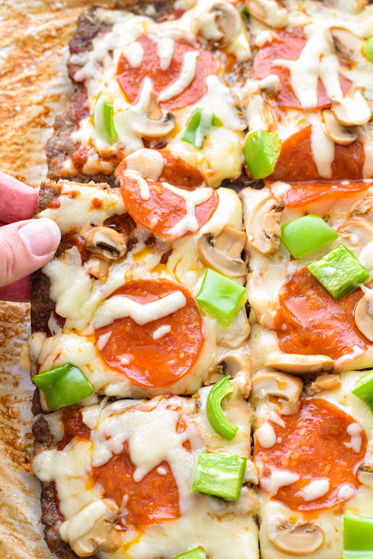 a hand holding the corner of a piece of low fodmap keto meatza with cheese, pepperoni, mushrooms and green peppers