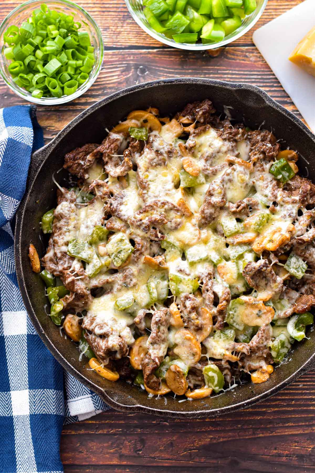 overhead shot of low fodmap keto philly cheesesteak skillet next to a towel and bowls of chopped scallions and green bell pepper