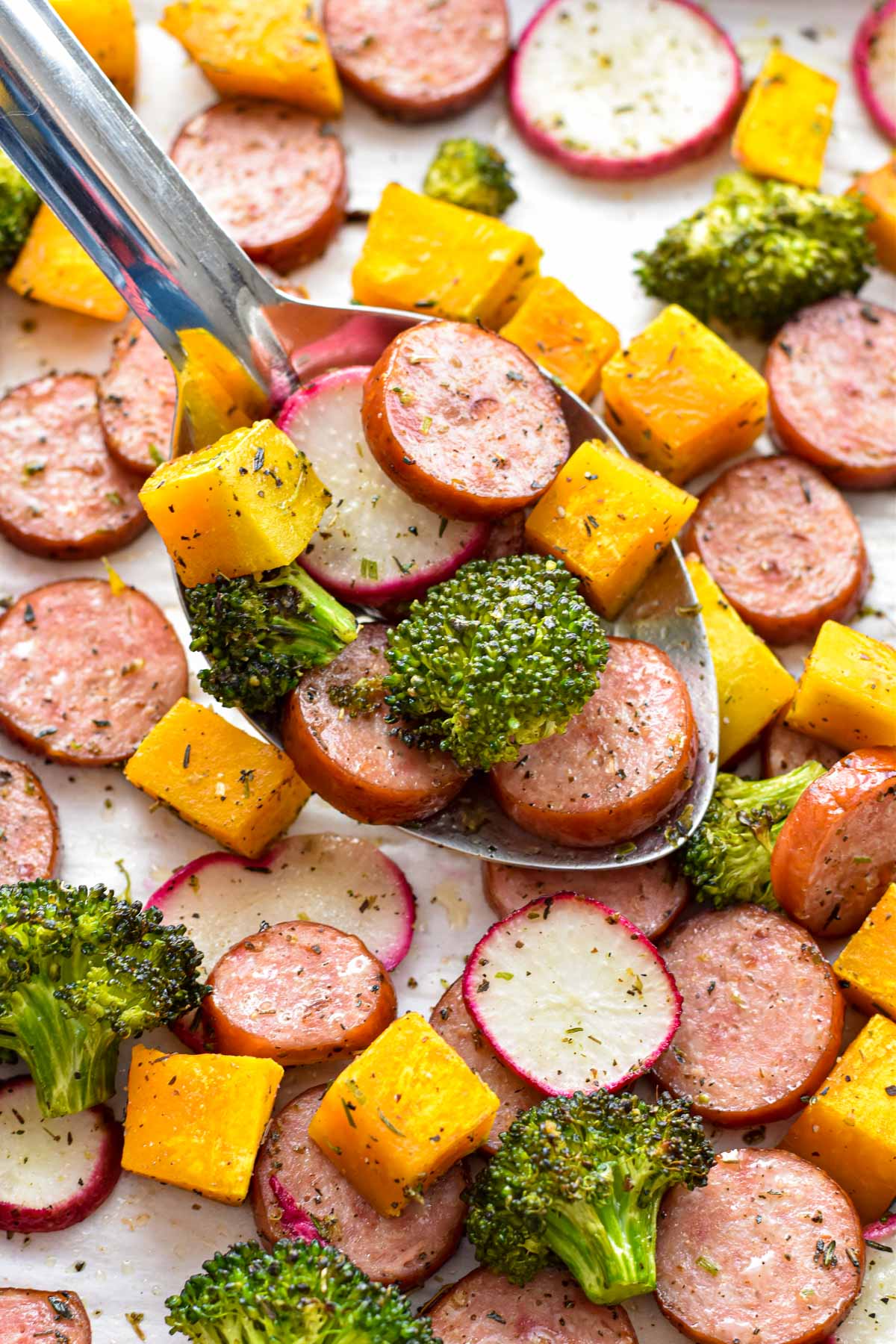 a spoon scooping low fodmap sausage and fall vegetables from a sheet pan covered in parchment paper