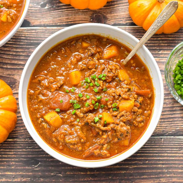 low fodmap pumpkin chili in a white bowl with a spoon next to orange gourds on a wooden background