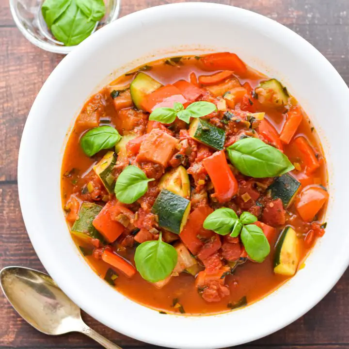 Low FODMAP ratatouille vegetable stew in a white bowl with tomatoes, red peppers, zucchini and basil.