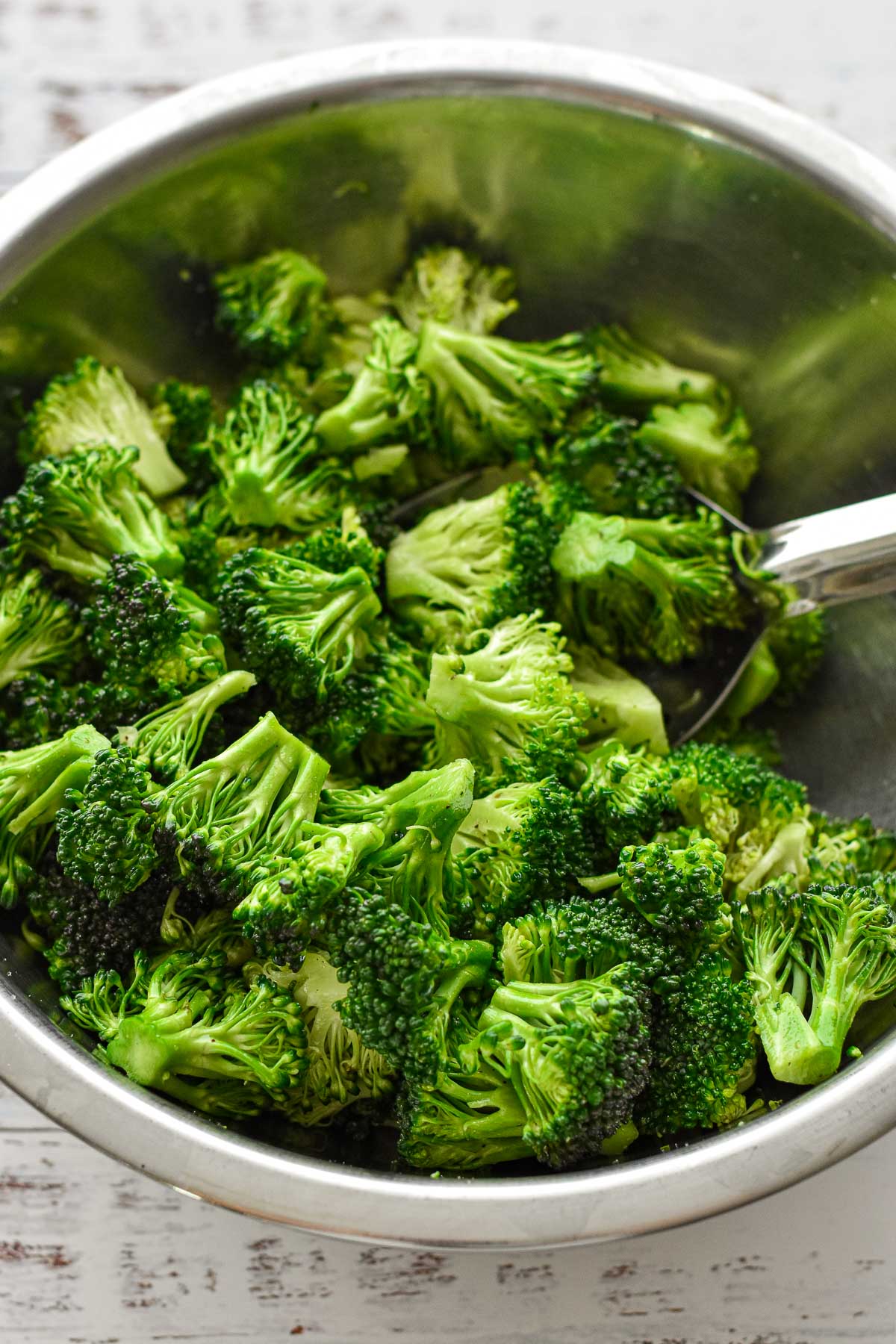 process shot of broccoli in a stainless steel bowl after tossing with a spoon to coat in oil mixture