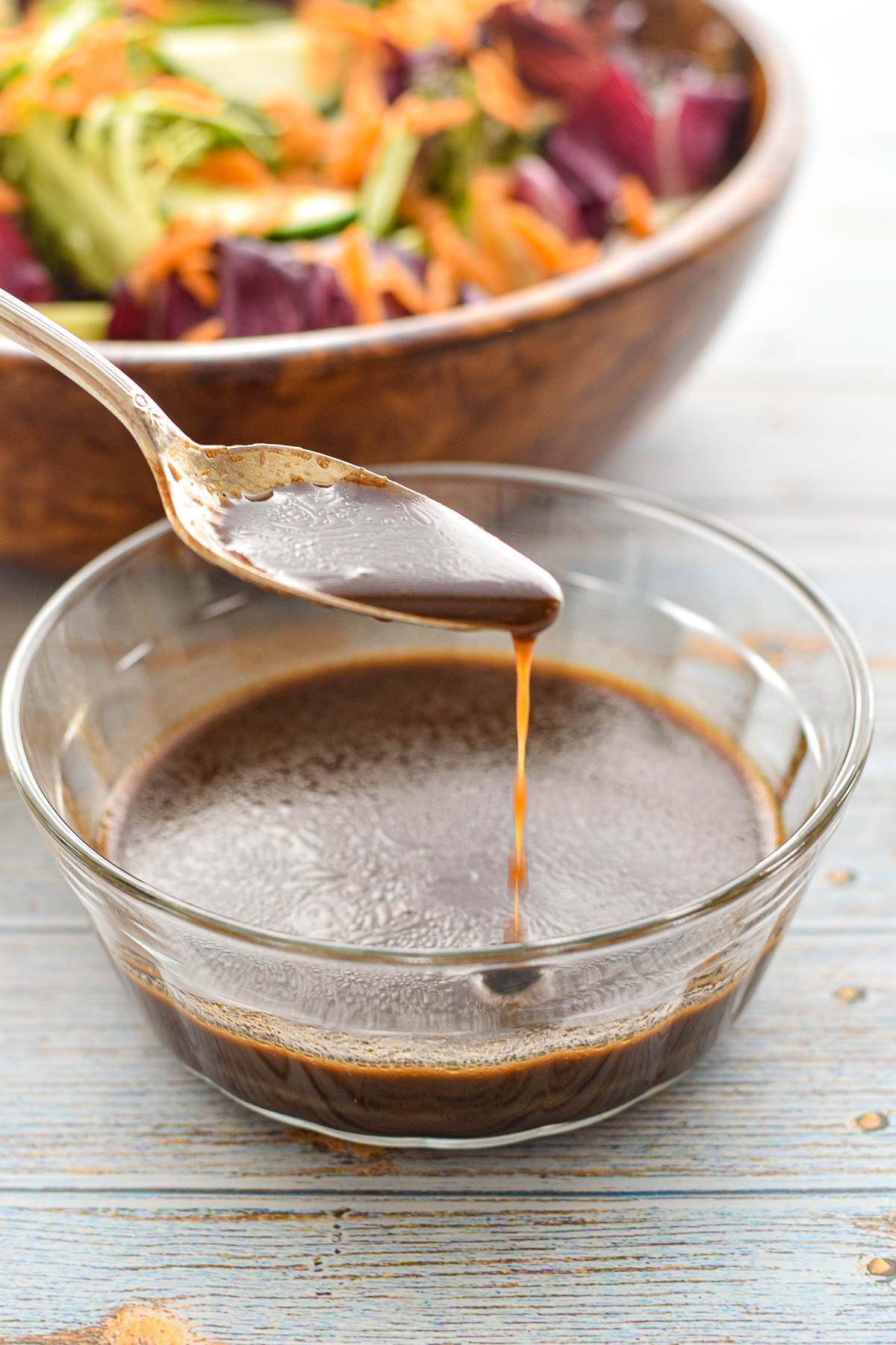 a spoon pouring balsamic vinaigrette into a glass bowl with a salad behind