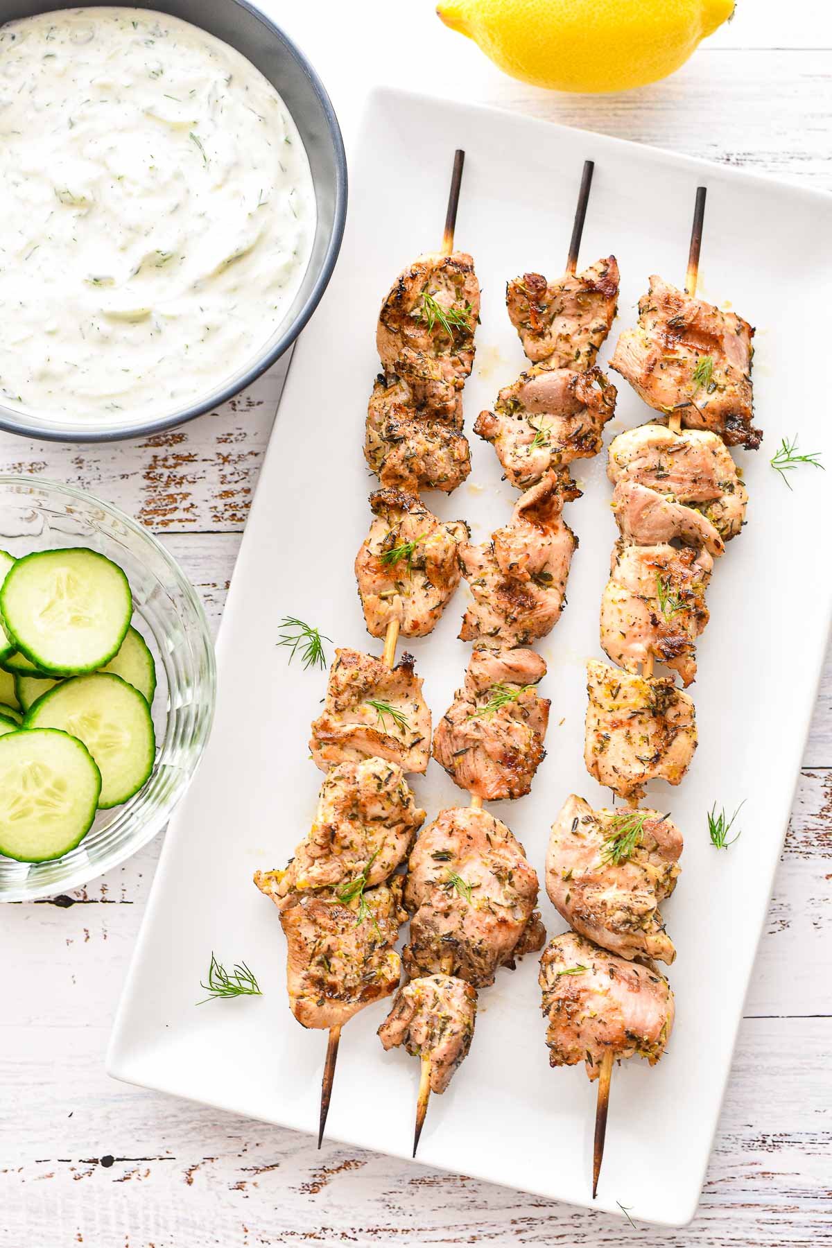 overhead shot of low fodmap greek chicken souvlaki garnished with fresh dill next to bowls of cucumber slices, tzatziki sauce, and a lemon