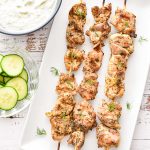low fodmap greek chicken souvlaki on wooden skewers on a white plate next to cucumber slices and a bowl of tzatziki sauce