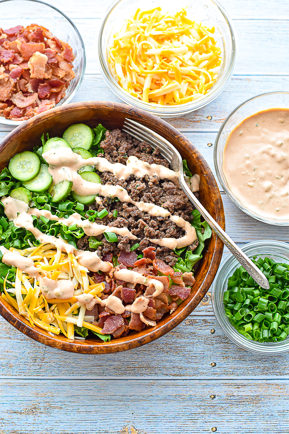 overhead shot of low fodmap burger bowl in a wooden bowl with a fork next to bowls of burger sauce, chopped scallions, shredded marble cheese, and chopped cooked bacon