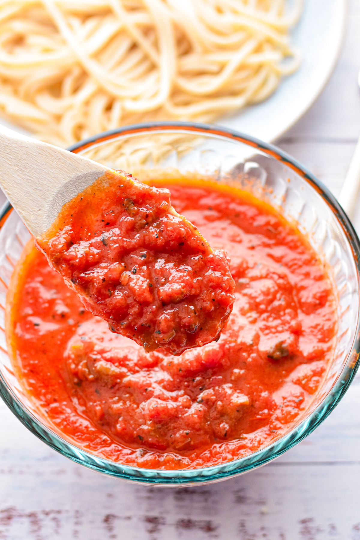 a wooden spoon covered in low fodmap spaghetti sauce over a bowl of sauce in front of a plate of low FODMAP spaghetti.