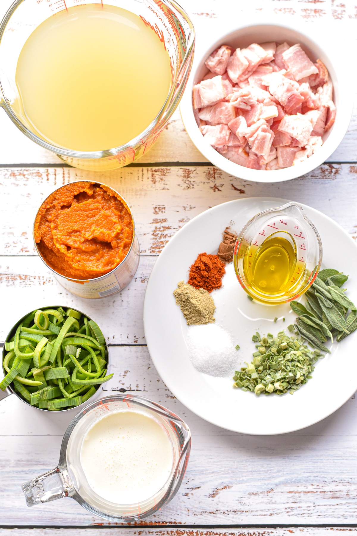 overhead shot of low fodmap pumpkin soup ingredients including chopped bacon, chicken bone broth, canned pumpkin puree, sliced leek greens, garlic-infused olive oil, fresh sage leaves, dried chives, salt, white pepper, smoked paprika, pumpkin pie spice, and heavy cream on a white background.