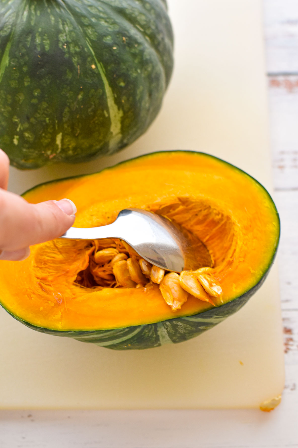 a hand scooping the seeds out of a half of a kabocha squash / japanese pumpkin with a dinner spoon on a cutting board.