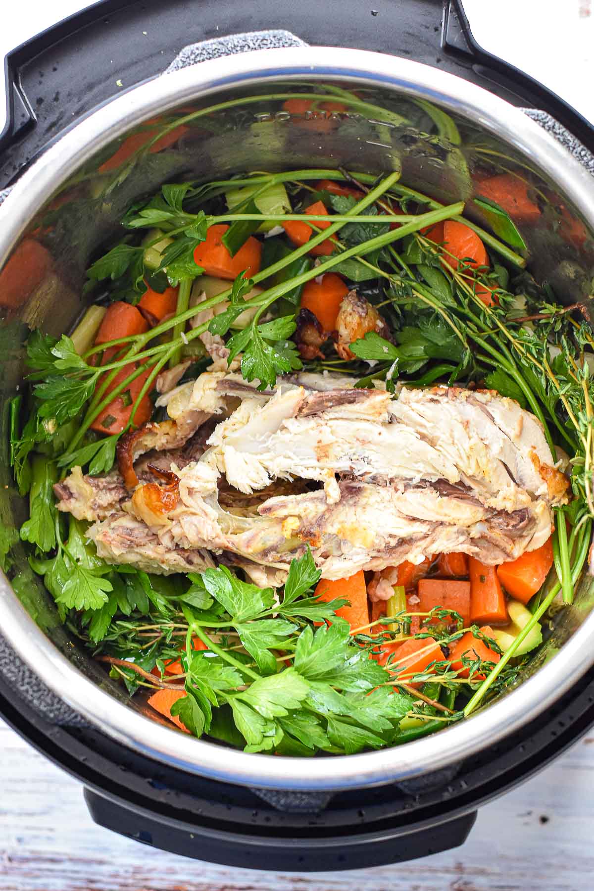a chicken carcass surrounded by parsley and thyme sprigs on top of sauteed veggies in an instant pot prior to being covered with water and cooking.