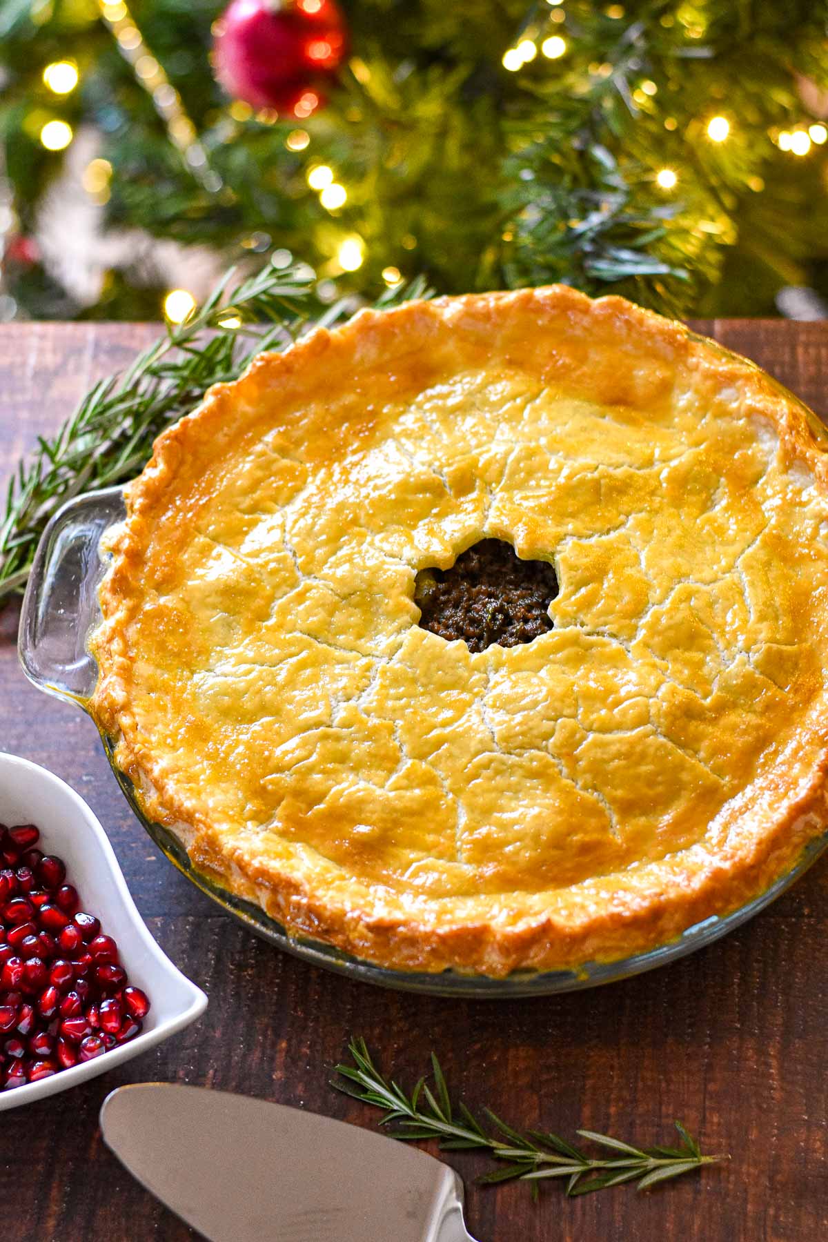 low fodmap tourtiere with a gluten-free crust next to rosemary sprigs and a bowl of pomegranate arils on a table in front of a christmas tree.