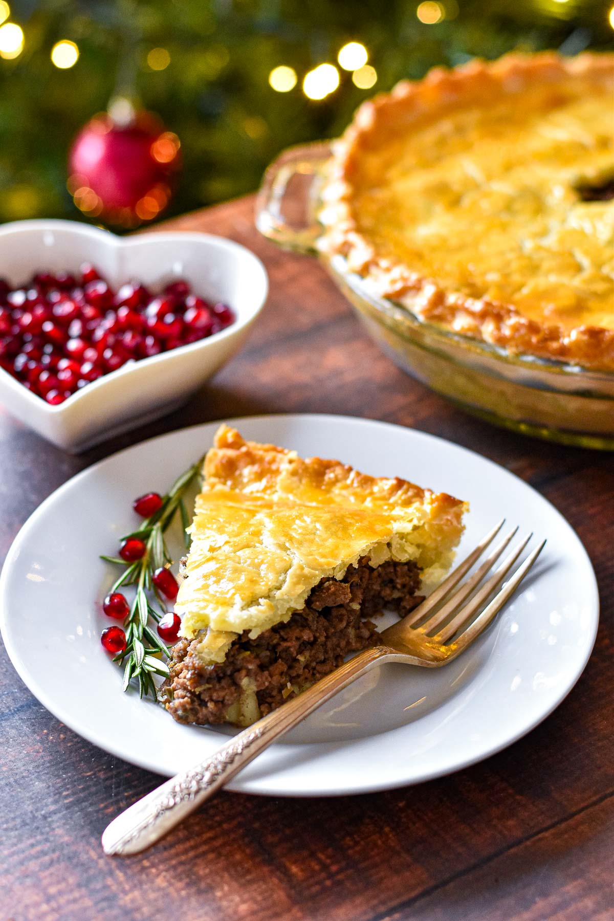 a slice of low fodmap tourtiere on a plate with a fork, rosemary sprig, and pomegranate seeds in front of a tourtiere and a bowl of pomegranate seeds on a table in front of a christmas tree.