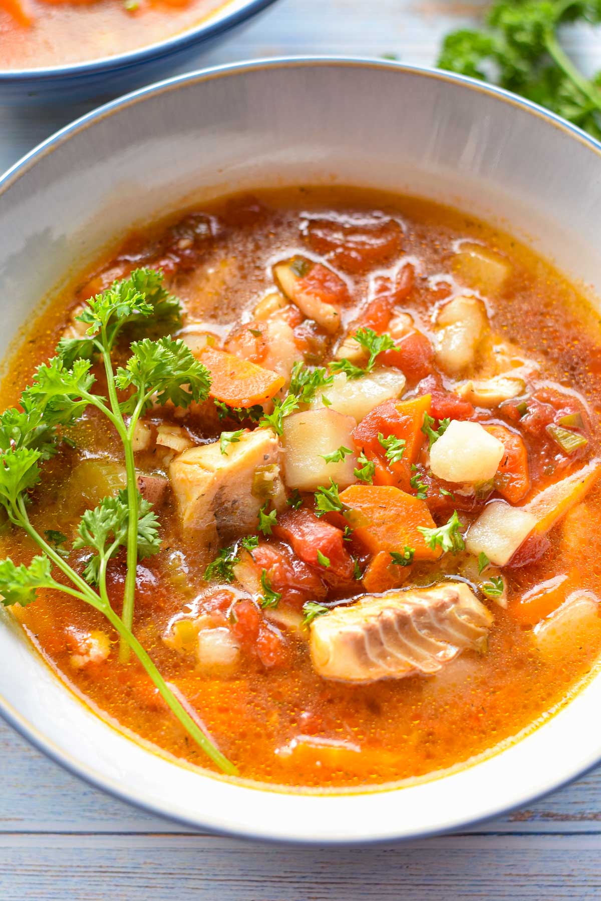 close up of Mediterranean style fish stew in a blue rimmed bowl with white fish, tomatoes, carrots, and potatoes.