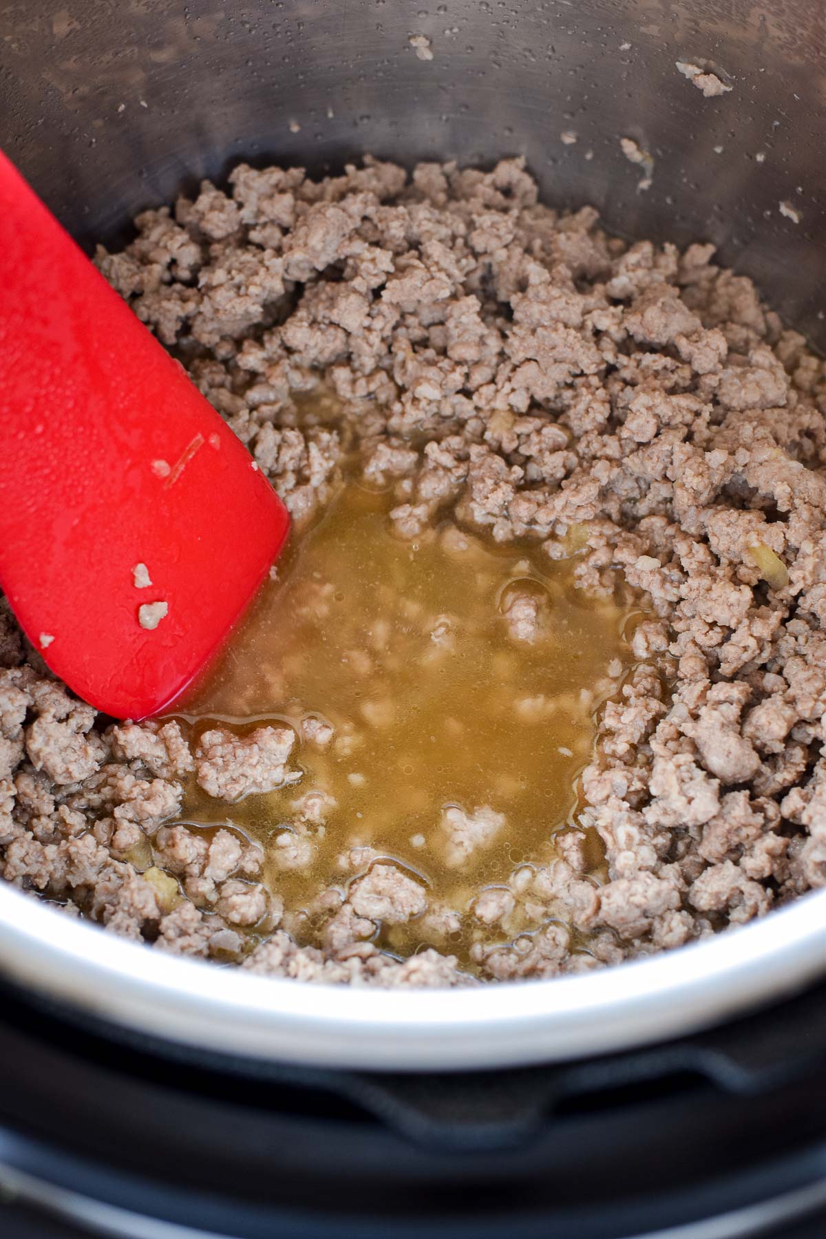 a spatula scraping the bottom of an instant pot clean using chicken bone broth after sautéing ground pork and ginger.